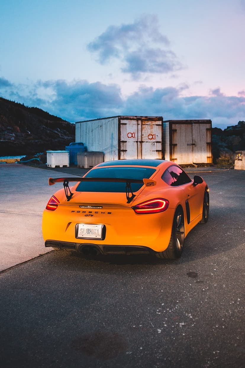 Porsche Cayman GT4 in orange with a rear wing parked on asphalt between shipping containers at sunset.