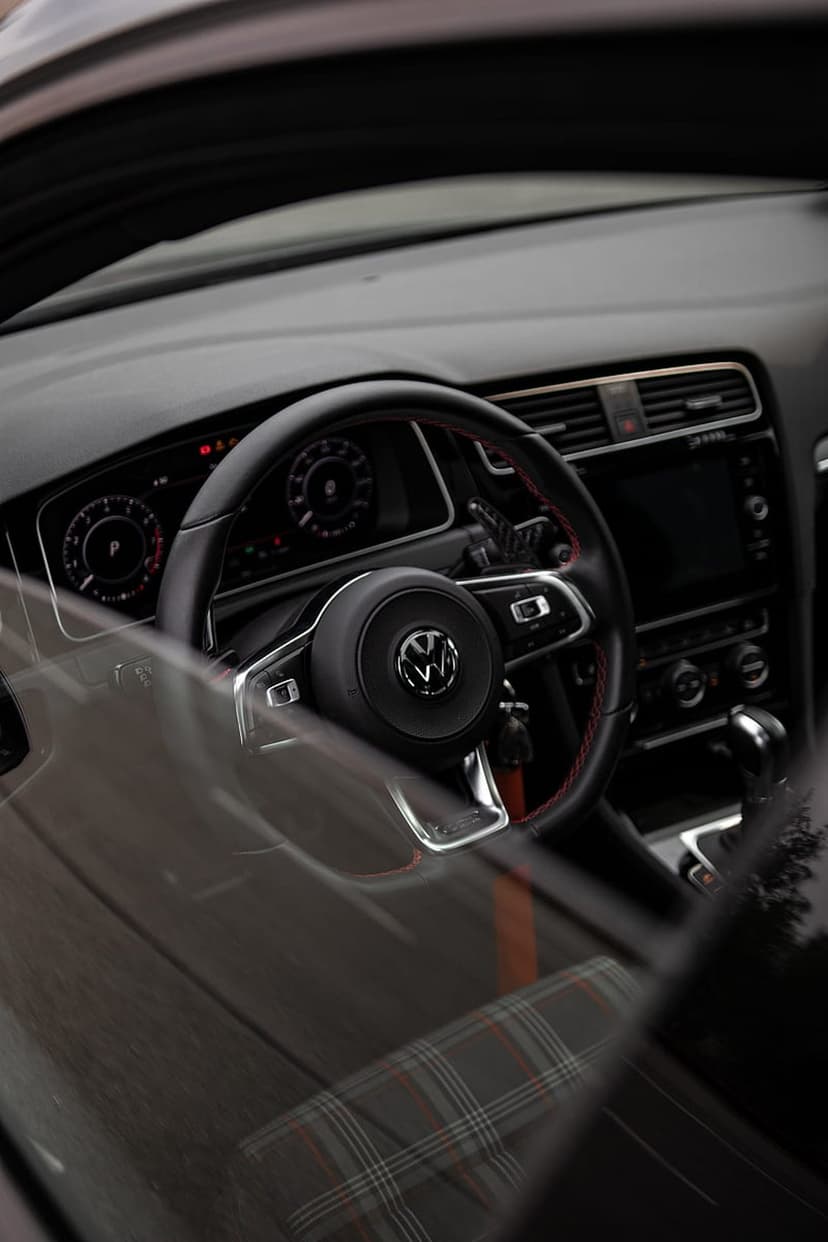 Volkswagen GTI steering wheel with red stitching, a digital dashboard, and tartan seat fabric in a car interior.