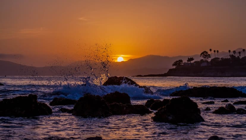 Orange sun dipping behind hills casts a warm glow over crashing ocean waves and dark jagged rocks along the coast.