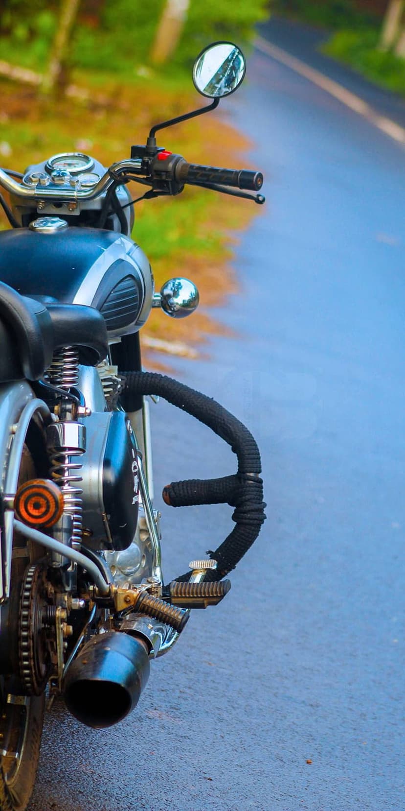 Vintage motorcycle handlebars and chrome engine components rest on a wet asphalt road with green foliage behind.