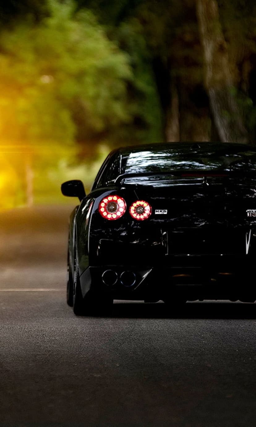 Nissan GT-R circular red taillights illuminated on a black car parked on a sun-dappled forest road at sunset.