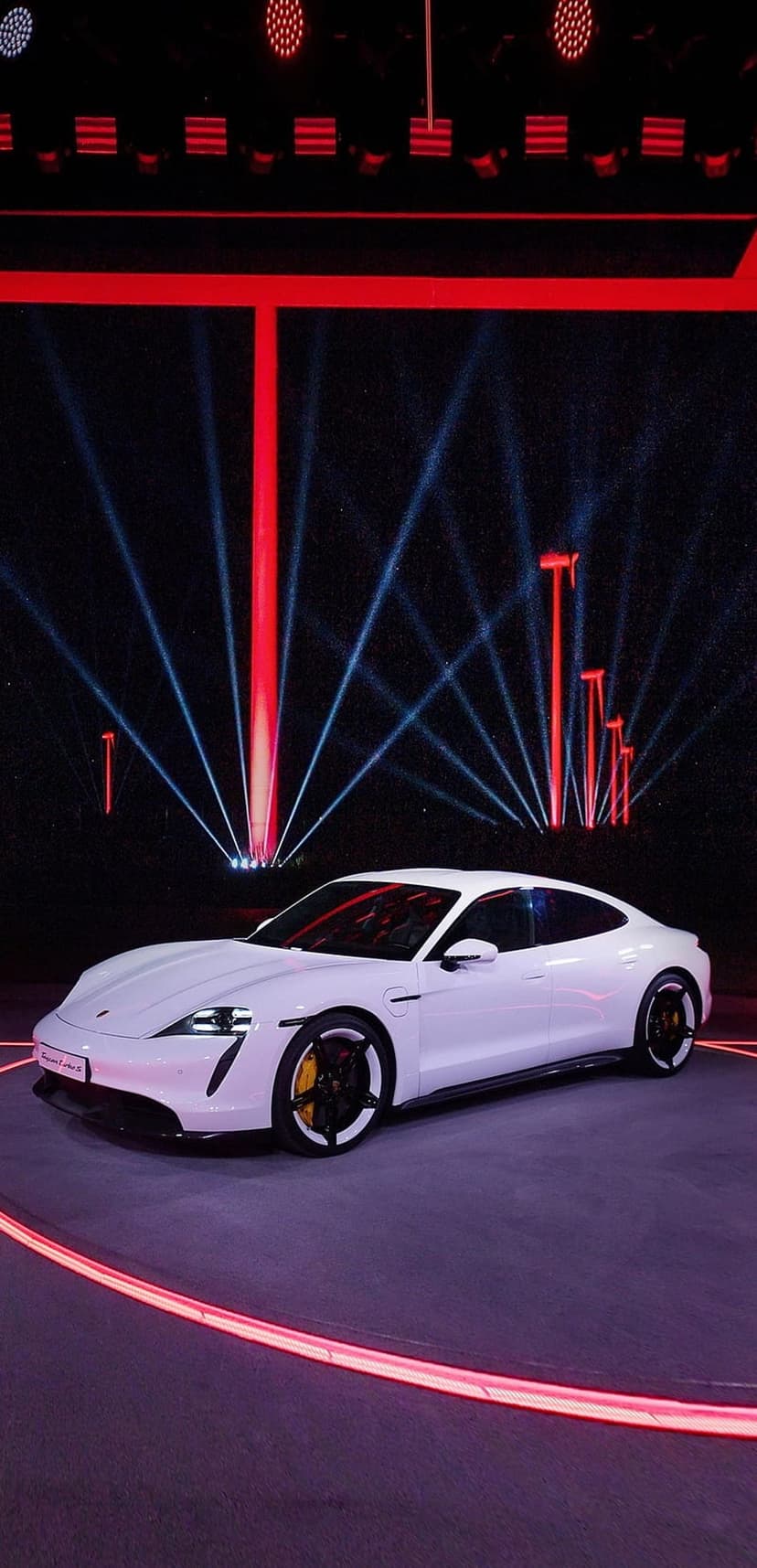 White Porsche Taycan electric car on a dark stage illuminated by vibrant red and blue neon spotlight beams.
