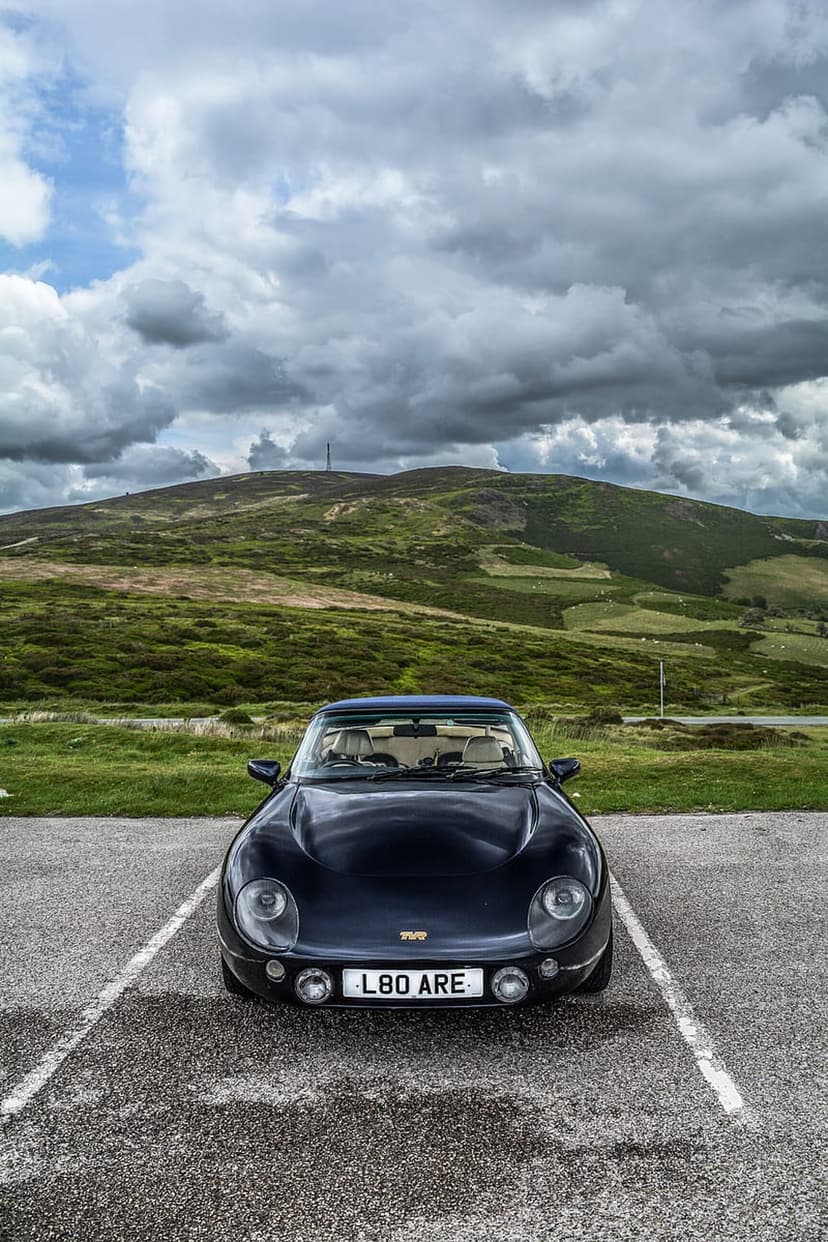 Dark TVR sports car parked on gravel with rolling green mountains and an overcast sky in the background.