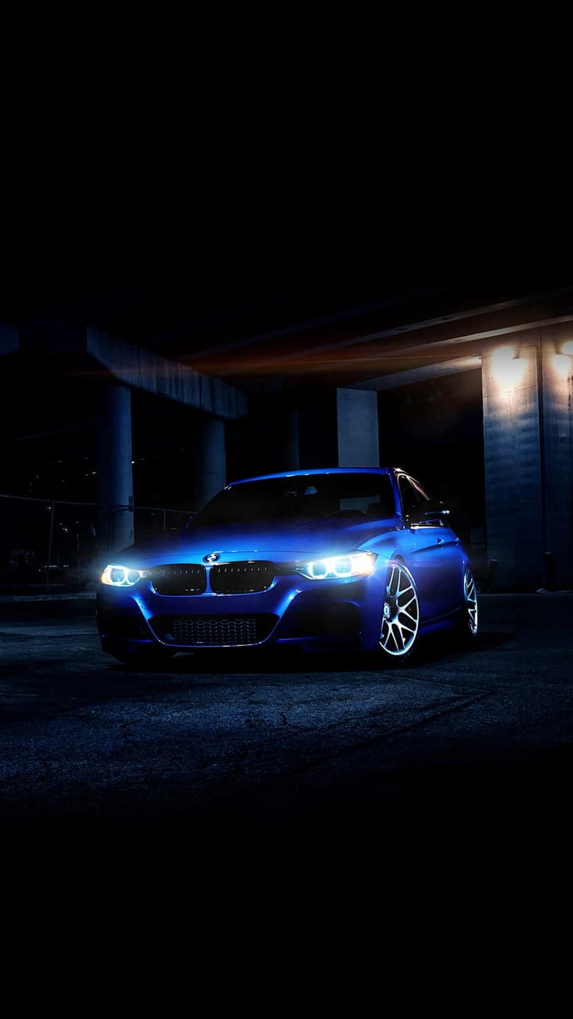 Blue BMW 3 Series with bright LED headlights parked under an urban overpass on dark asphalt during the night.