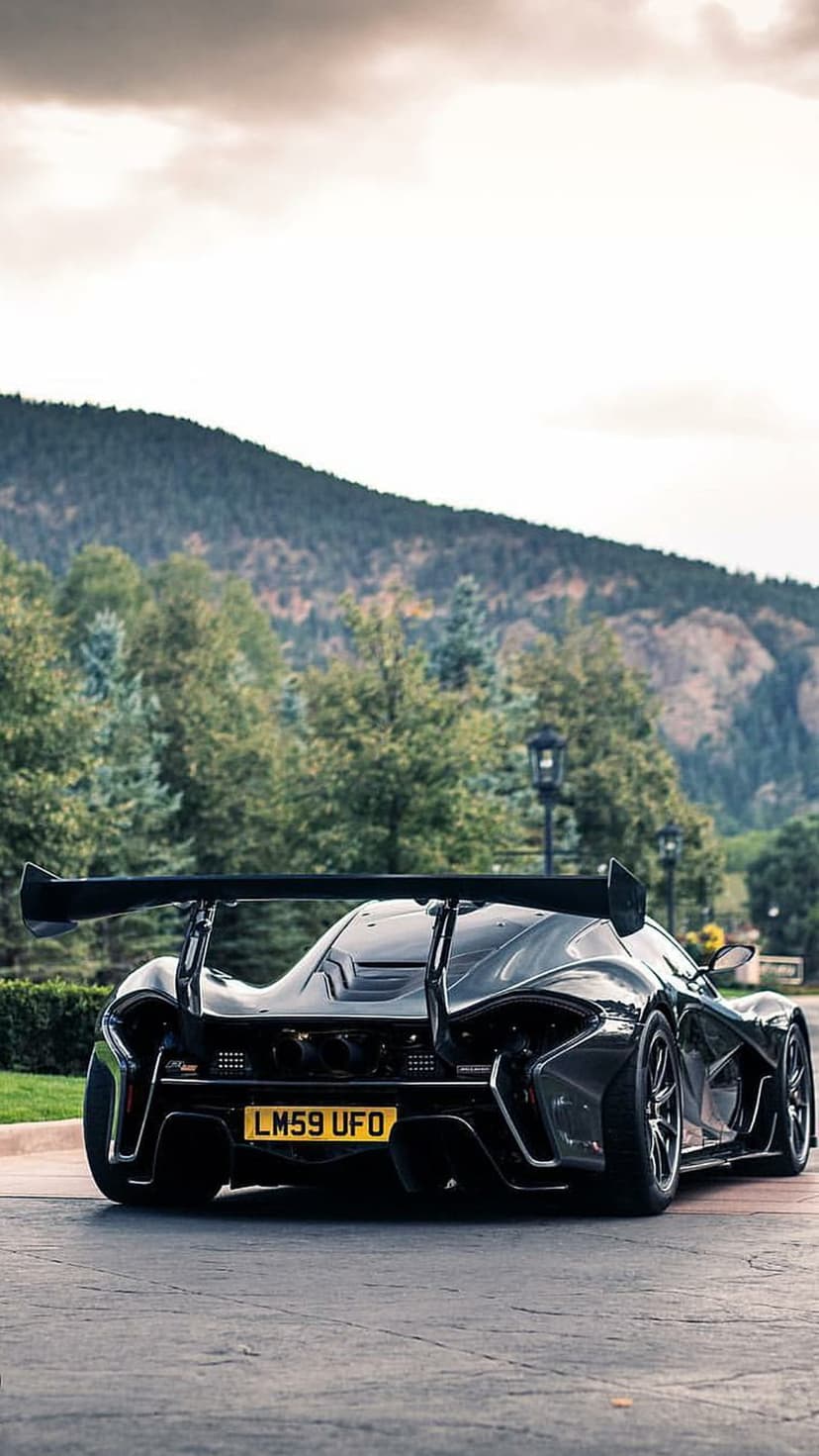 McLaren P1 GTR rear wing on a paved road with green mountains and a cloudy sky in a vertical car photography shot.