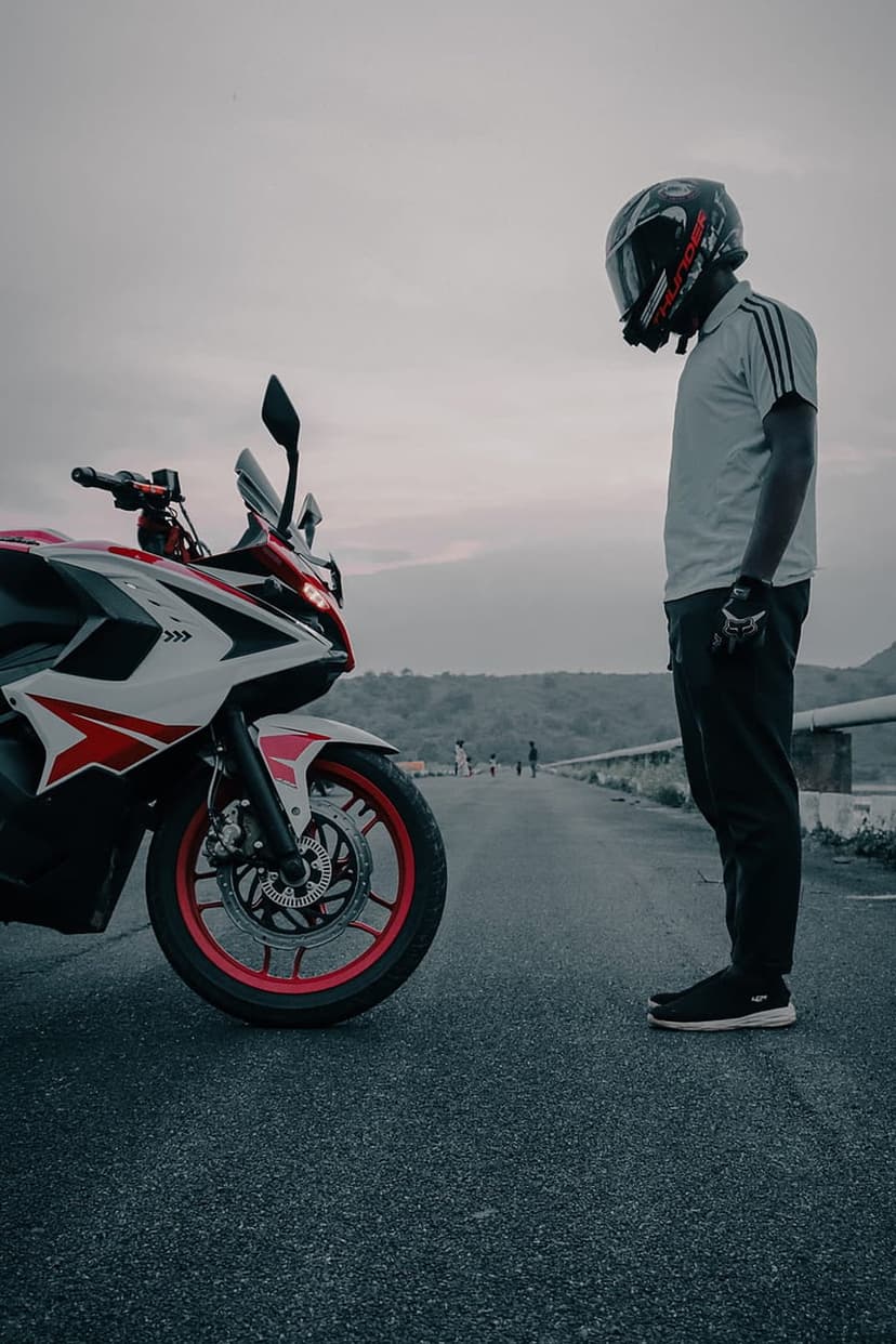 Helmeted rider standing beside a white sport motorcycle on an asphalt road under a cloudy, overcast sky.