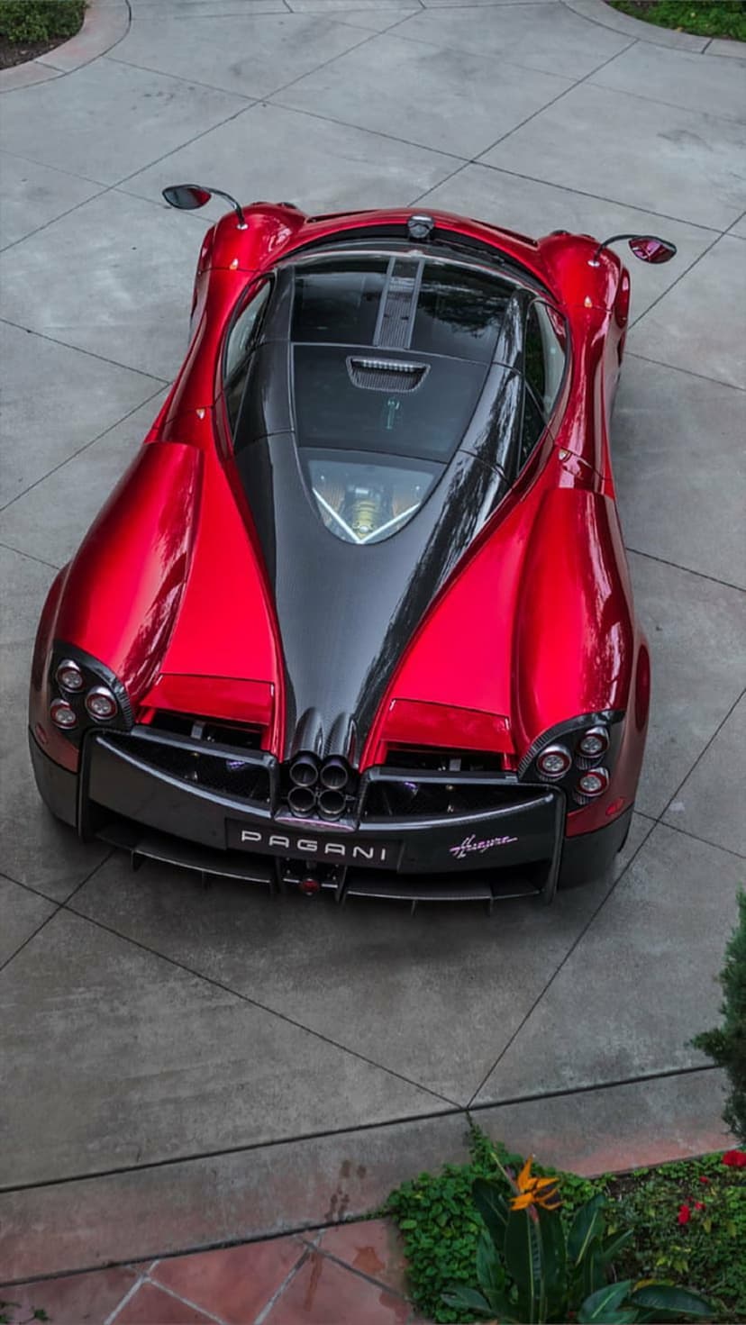 Red Pagani Huayra hypercar with a black center stripe parked on a driveway near green tropical foliage.