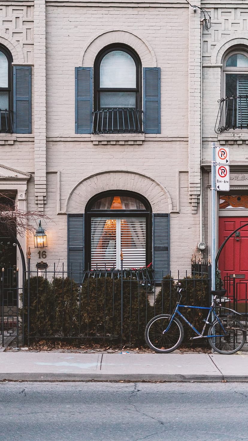 Blue bicycle parked by a red door and brick townhouse with blue shutters and a street lamp on a city sidewalk.