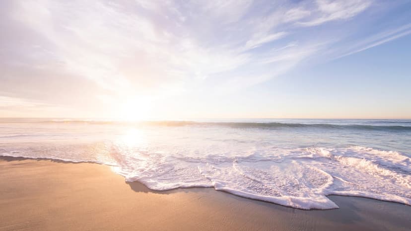 Golden sun rising over a calm ocean as thin waves reach a sandy shore under a sky filled with pastel clouds.