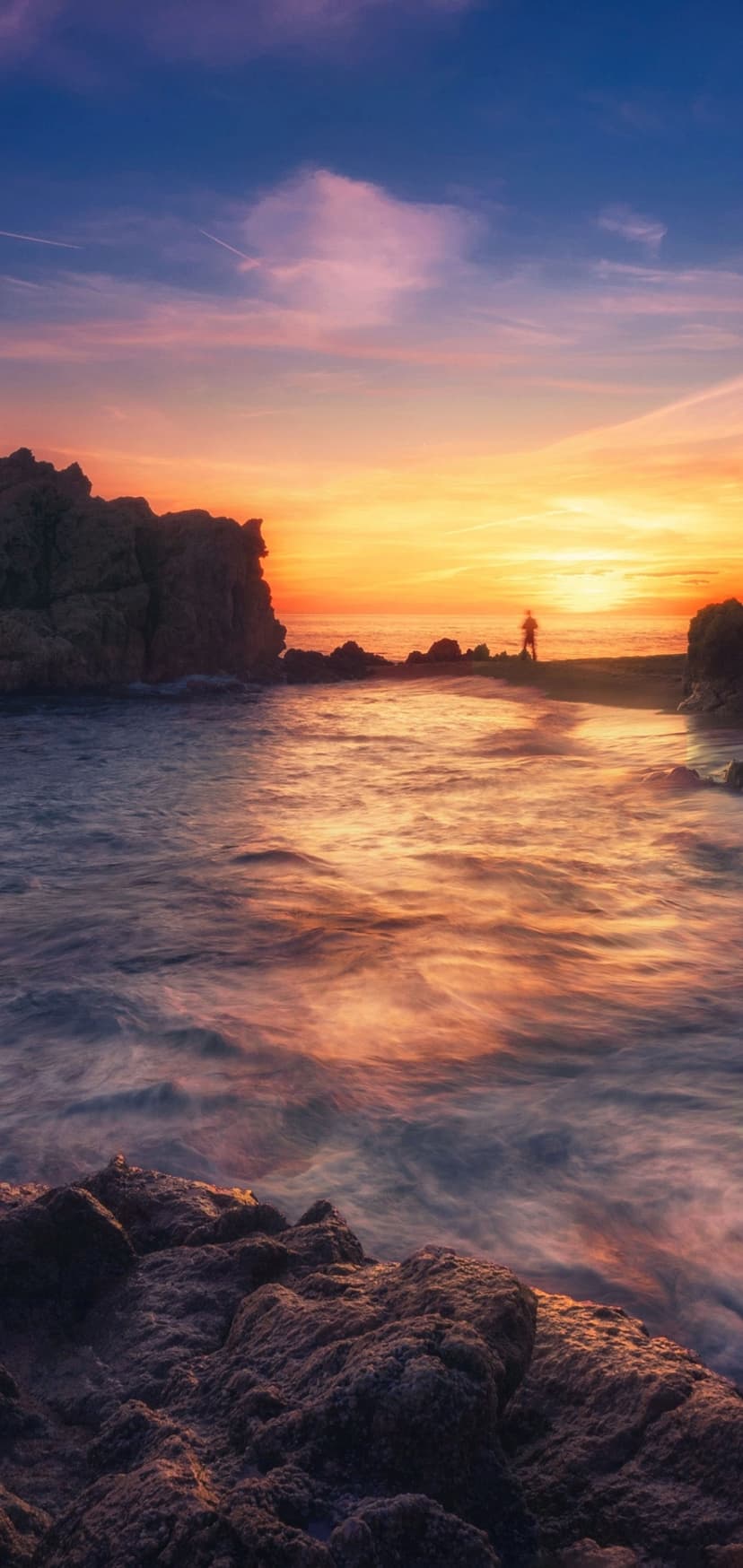 A lone silhouette stands on craggy rocks overlooking a vibrant pink and orange sunset over misty ocean waves.