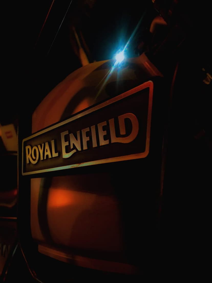 Royal Enfield chrome emblem glows under bright blue light with amber lens flares against a dark motorcycle frame.