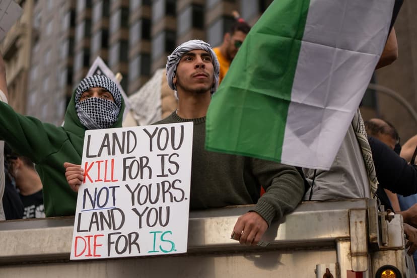 Protester in a keffiyeh holds a sign about land rights next to a waving Palestinian flag at a demonstration.
