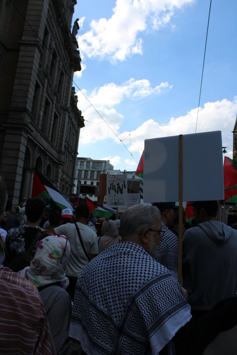 Palestinian flags held by a crowd wearing keffiyehs in a city street under a cloudy sky during a public rally.