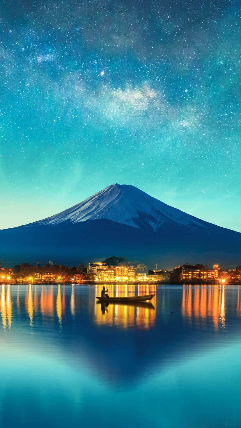 Mount Fuji rises behind a glowing lakeside town and a wooden boat under a starry night sky with the Milky Way.