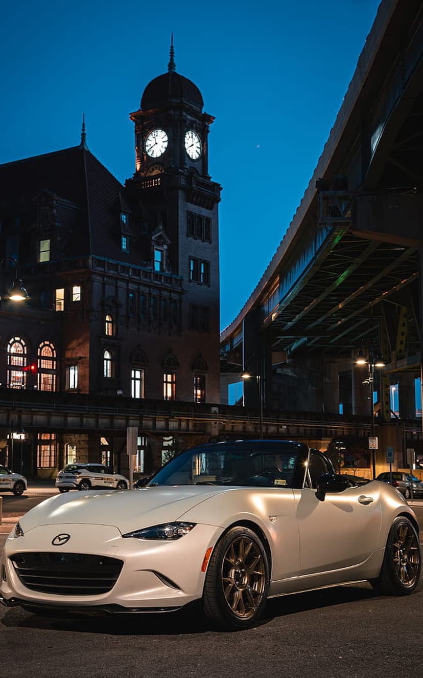 White Mazda Miata convertible with illuminated headlights parked on a city street near a historic clock tower.