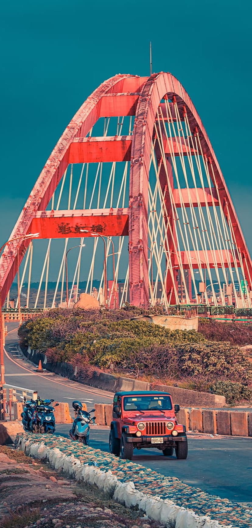 Red arch bridge with white cables towers over a coastal road featuring a red Jeep and green shrubbery.