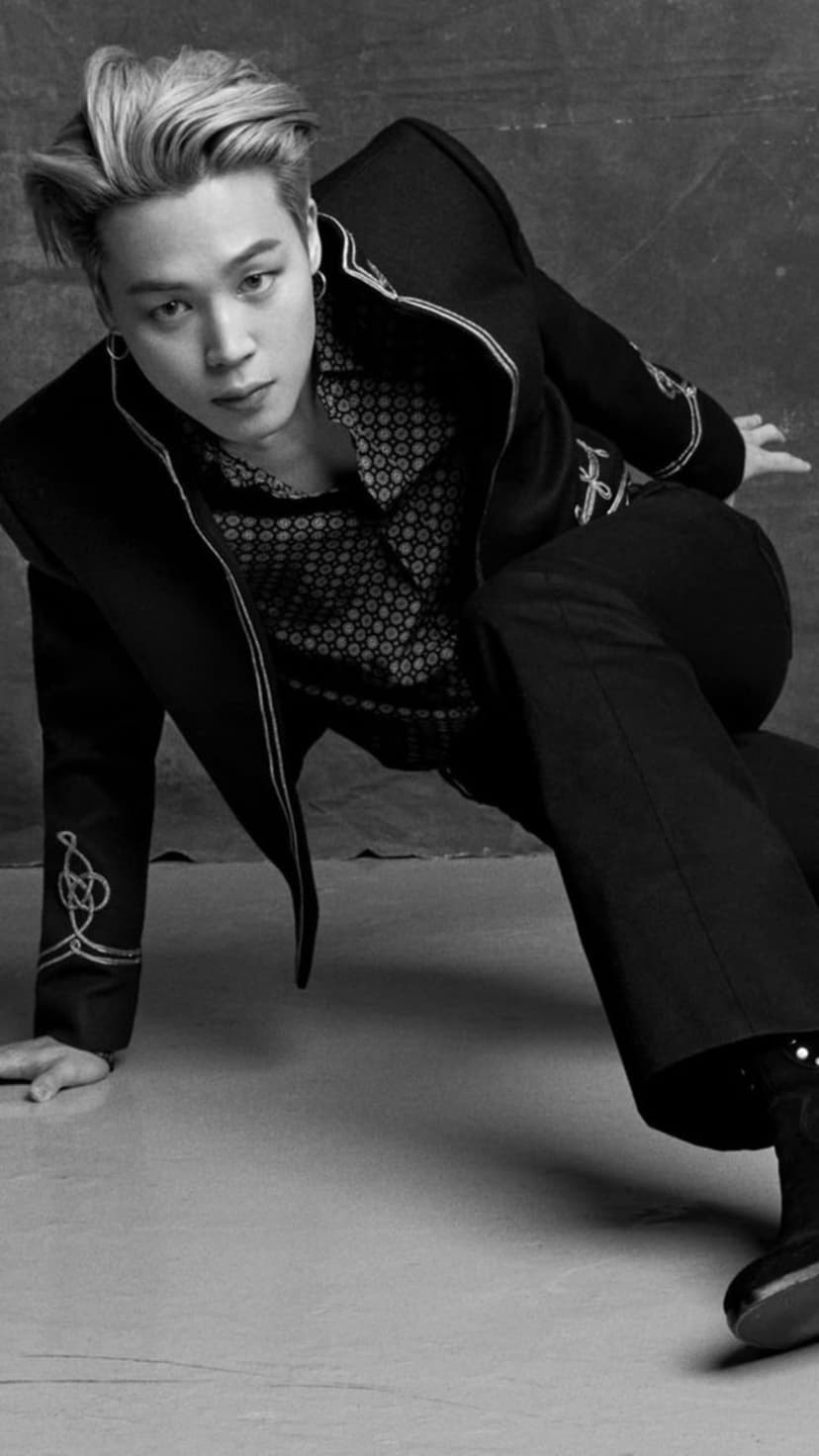 Jimin of BTS in a patterned shirt and embroidered jacket posing for a black and white dramatic low-angle portrait.