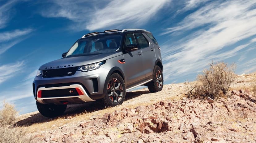 Grey Land Rover Discovery SVX SUV parked on a steep rocky hill against a bright blue sky with wispy clouds.