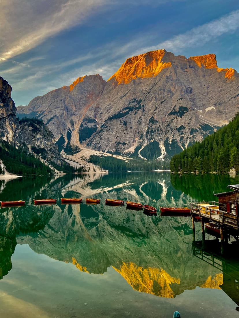 Wooden rowing boats anchor on a glassy emerald lake reflecting jagged sunlit mountain peaks and pine forests.