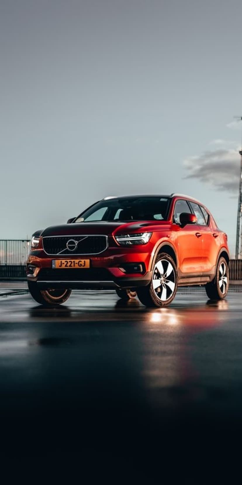 Red Volvo XC40 SUV parked on a wet street reflecting a golden sunset sky with distant clouds and a tower.