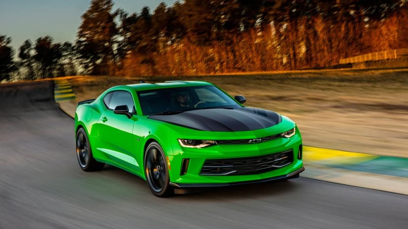 Green Chevrolet Camaro sports car with a black hood speeding along a racetrack with motion-blurred trees.