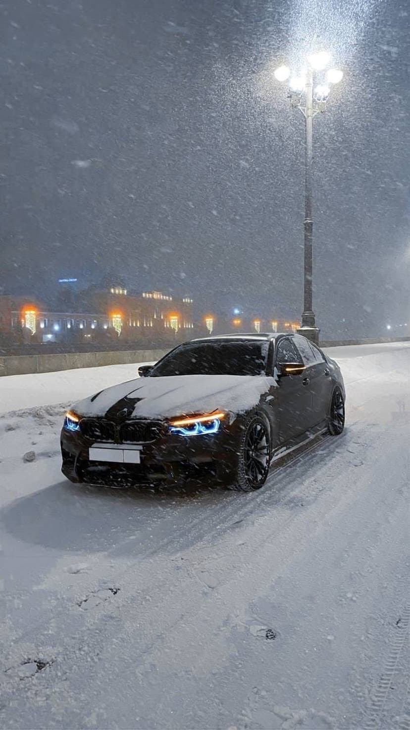 Luxury dark sedan parked on a snowy road at night with glowing headlights and falling snow under streetlights.