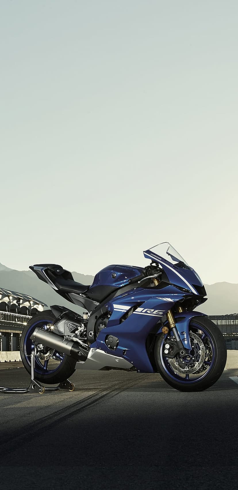 Blue Yamaha R6 sportbike on a paved racetrack with rugged mountains and a bright sky in the background.