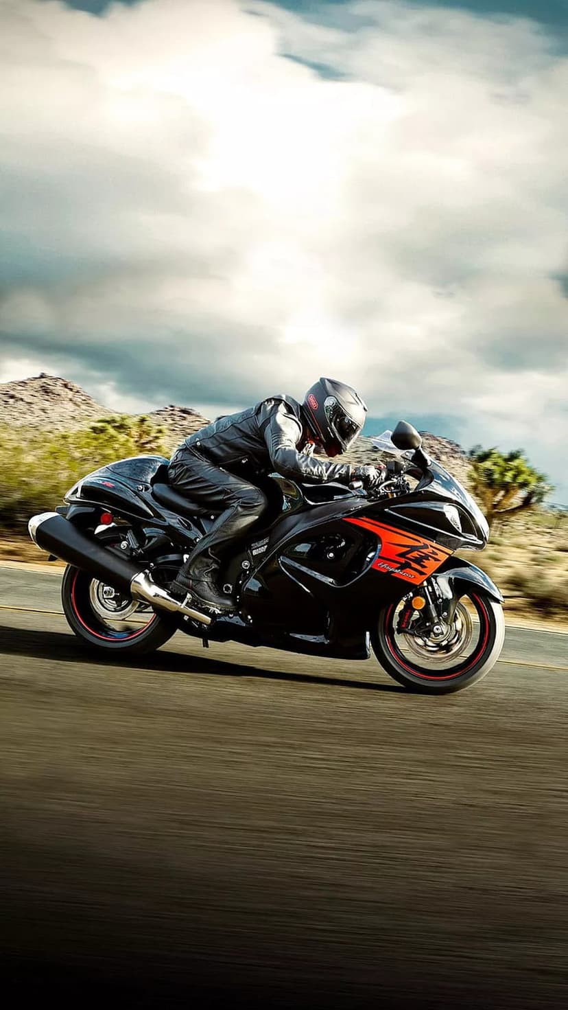 Motorcyclist on a black and orange sportbike speeds past desert mountains under a cloudy sky on a paved road.