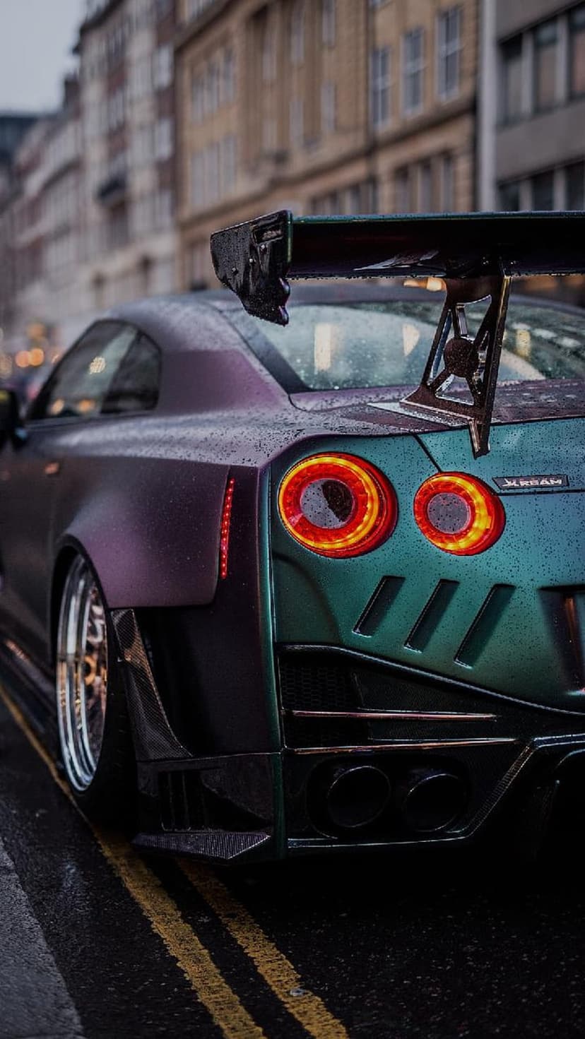Nissan GT-R with a widebody kit and rear spoiler parked on wet urban pavement under dim nighttime street lights.
