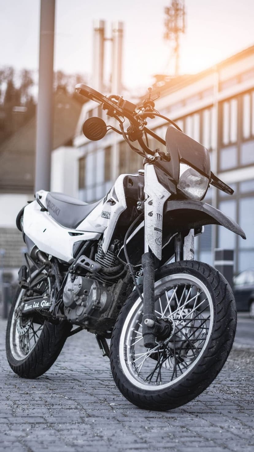 Suzuki dual-sport motorcycle with spoked wheels parked on a sunlit cobblestone street in an urban setting.