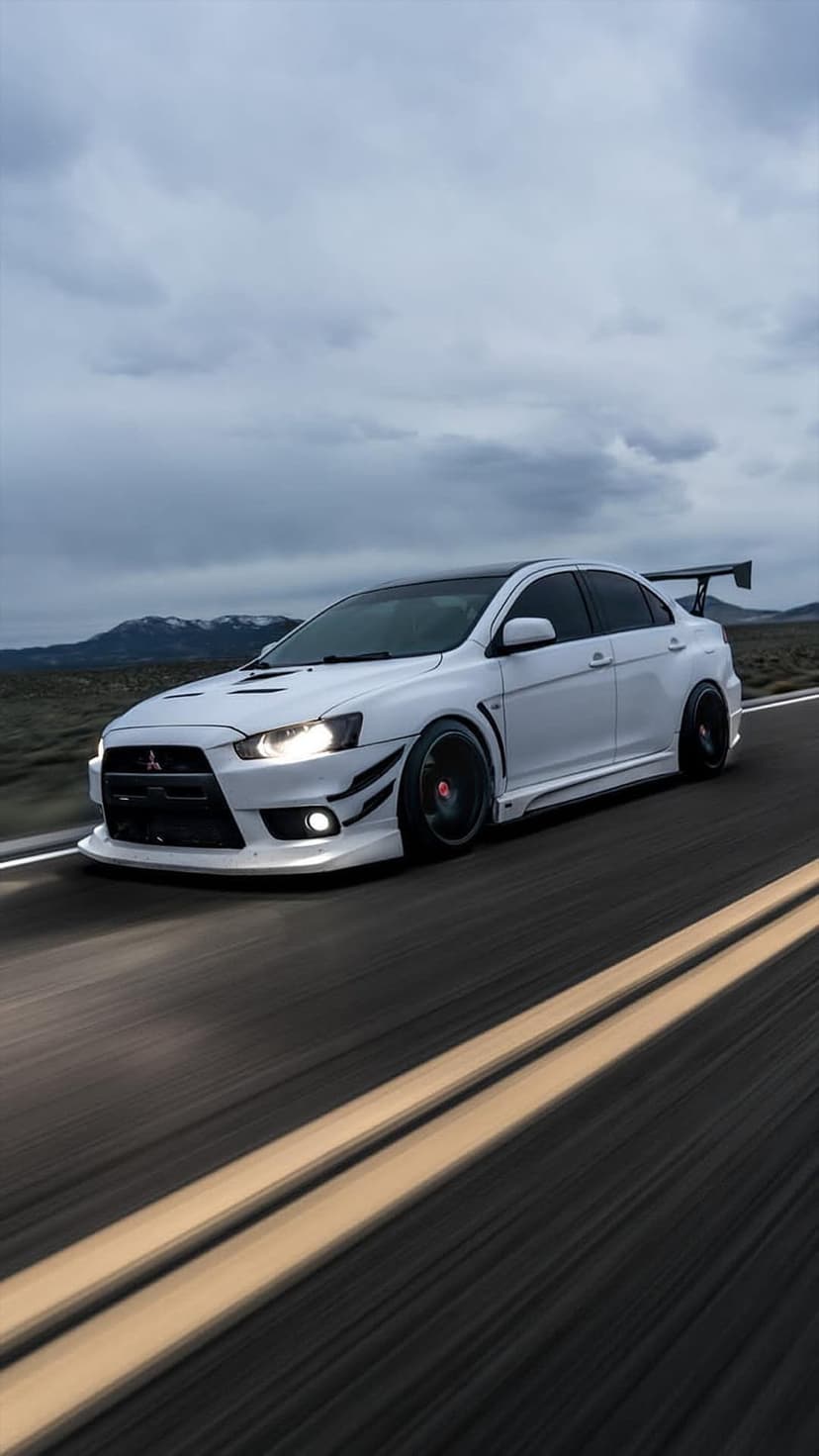 White Mitsubishi Lancer Evo X with a large rear wing drives along a winding road with distant mountain peaks.