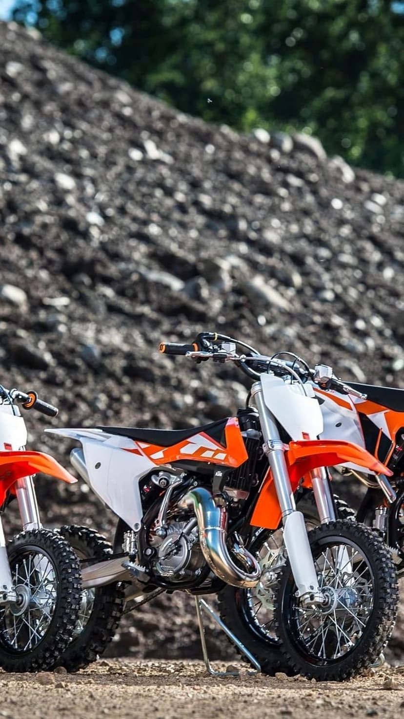 KTM dirt bikes with knobby tires parked on a dirt trail against a blurred backdrop of green trees and rocks.