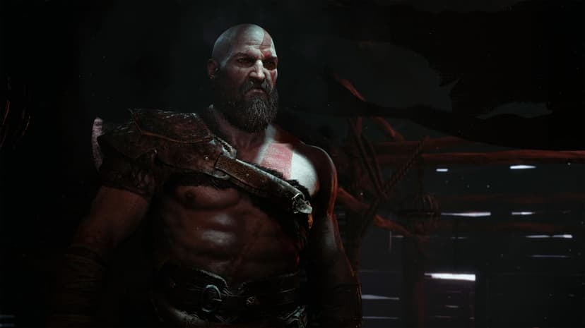 Kratos from God of War with a thick beard and red tattoos stands stoically in a dimly lit wooden interior.