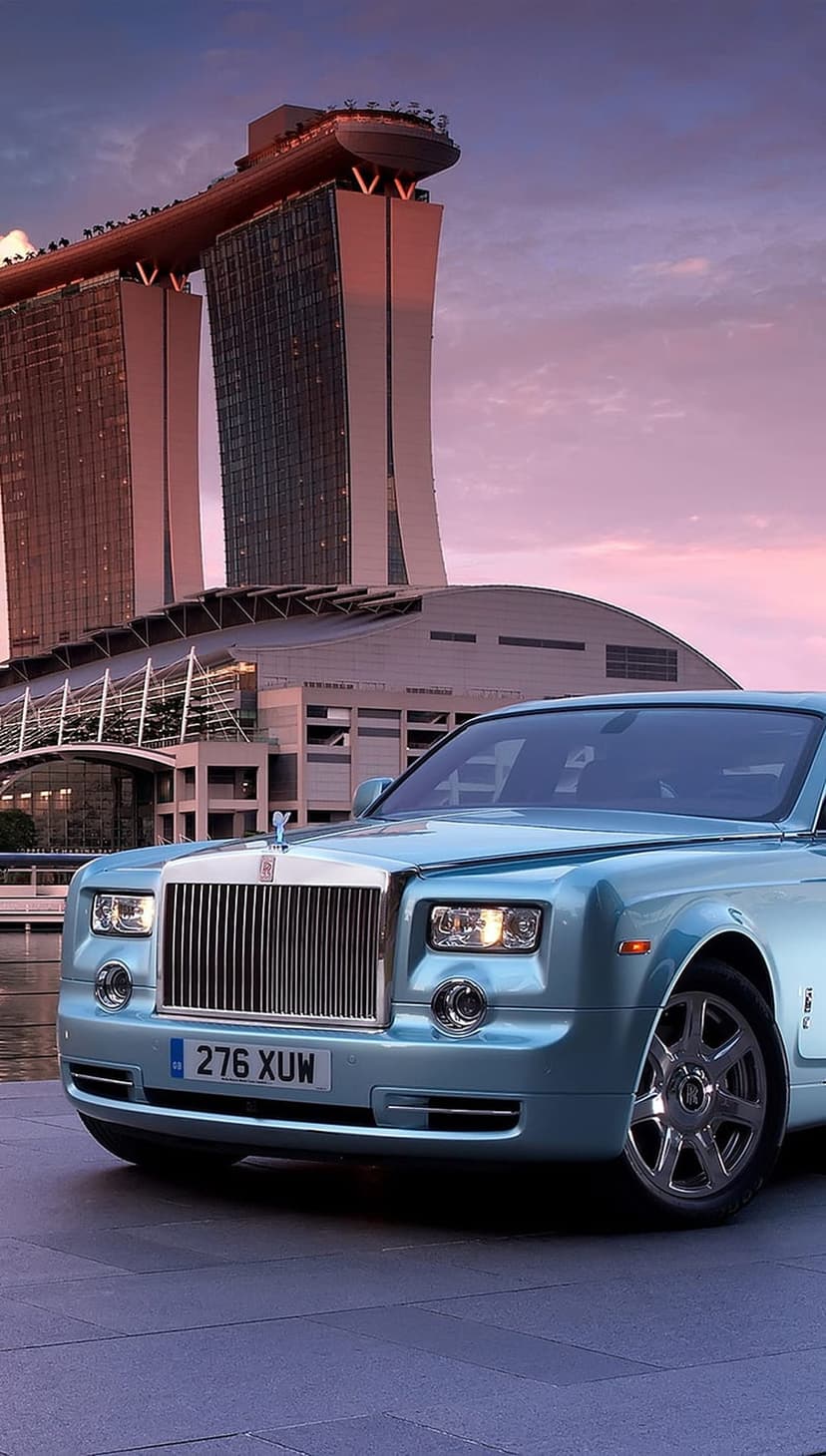 Light blue Rolls-Royce Phantom sedan parked in front of the Marina Bay Sands towers under a warm sunset sky.