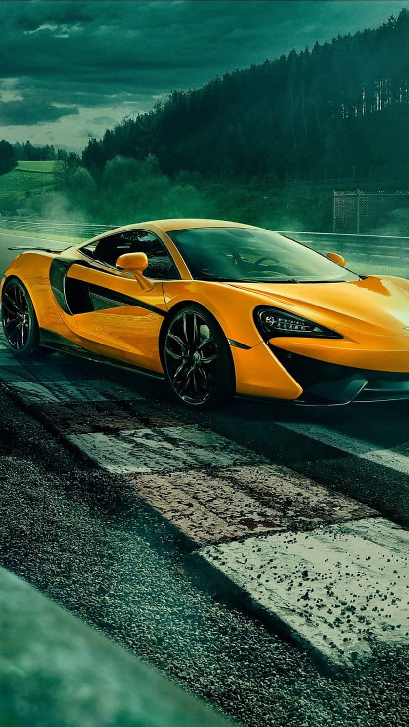 Orange McLaren supercar on a racing circuit with black rims, dark green hills, and a dramatic cloudy sky.