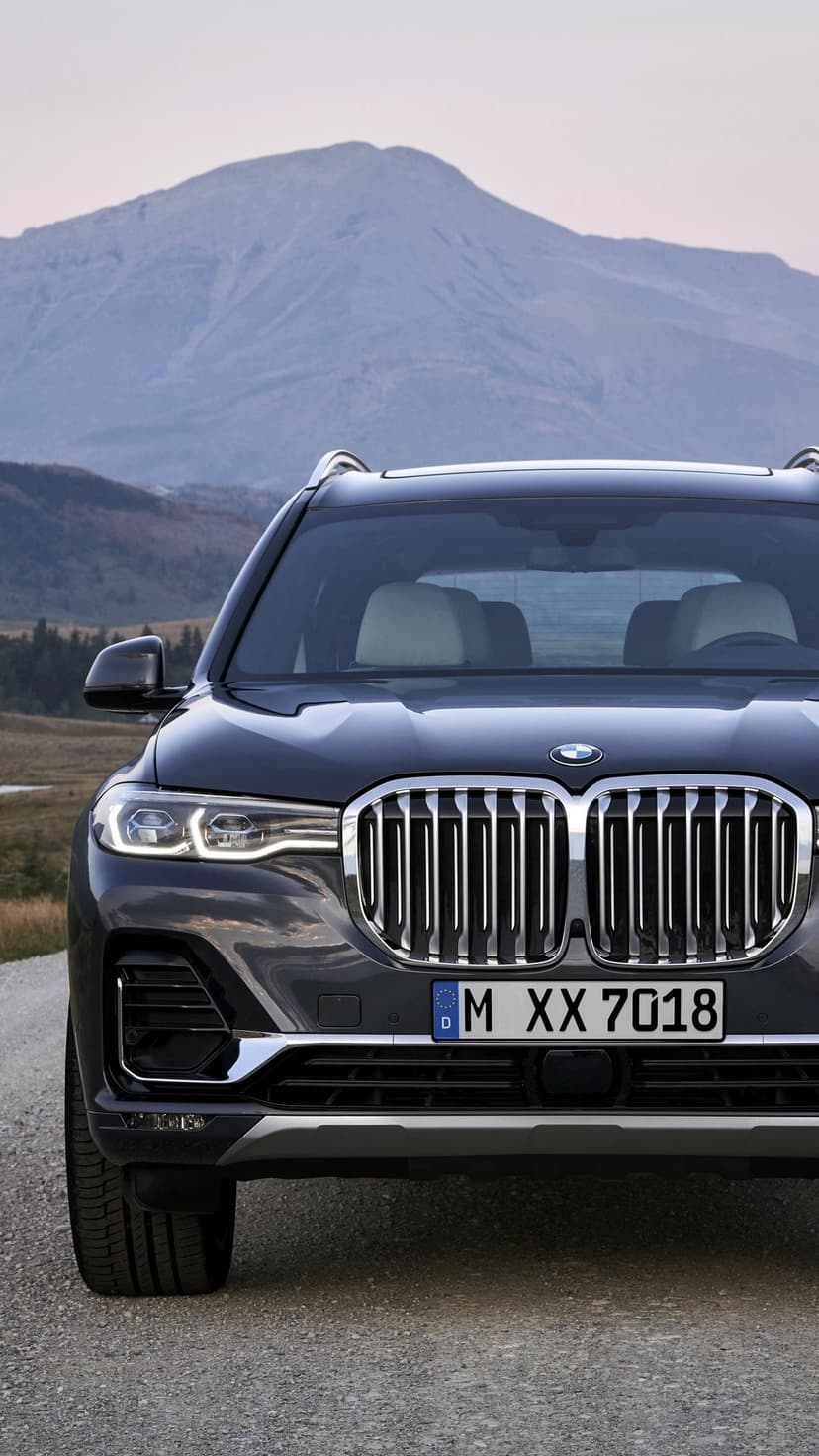 Dark grey BMW X7 SUV front view with chrome grille and LED lights against a hazy mountain background.