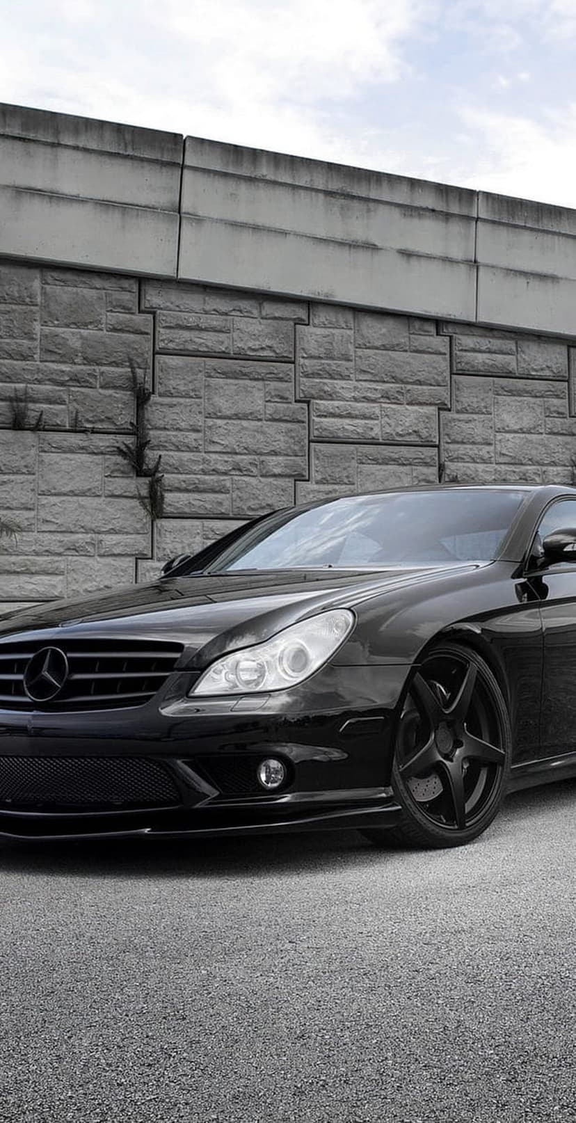 Black Mercedes CLS sedan parked at a low angle against a grey textured stone wall with black sport rims.