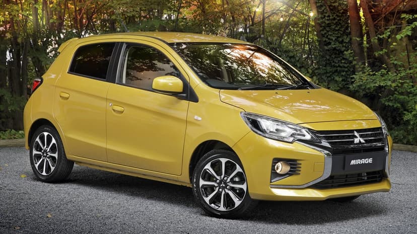Yellow Mitsubishi Mirage hatchback parked on gravel with sunlight filtering through lush green trees.