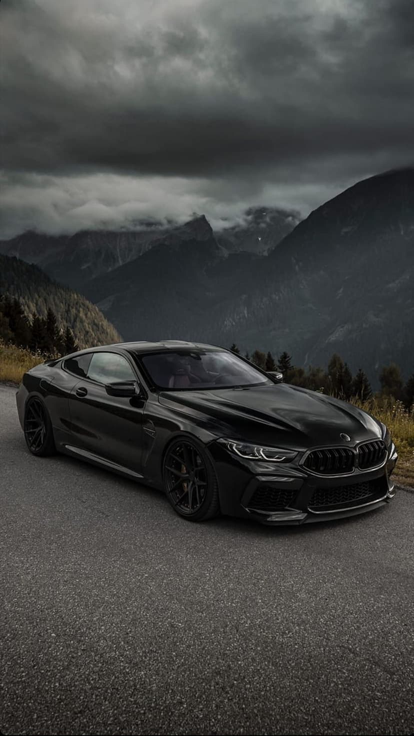 Black BMW M8 coupe parked on a mountain road with dark storm clouds and jagged peaks in the background.