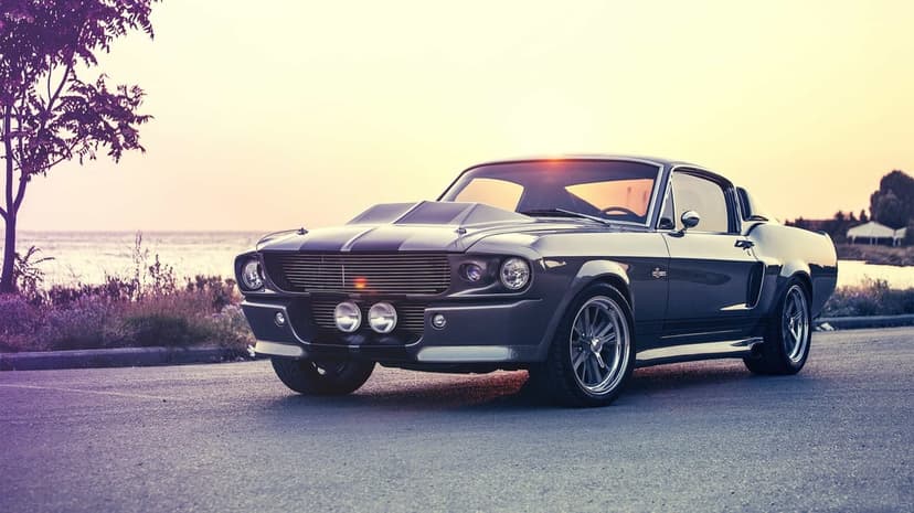 Charcoal grey classic muscle car on a coastal road at sunset with purple sky and distant water background.