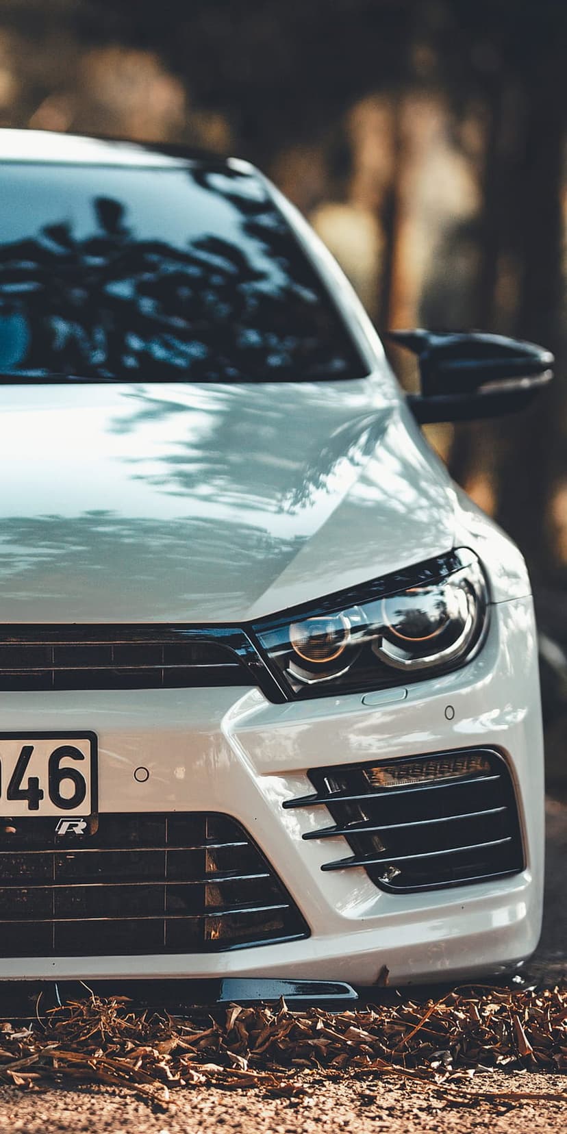 White Volkswagen Scirocco front bumper and headlights resting on dry orange autumn leaves in a blurred forest park.
