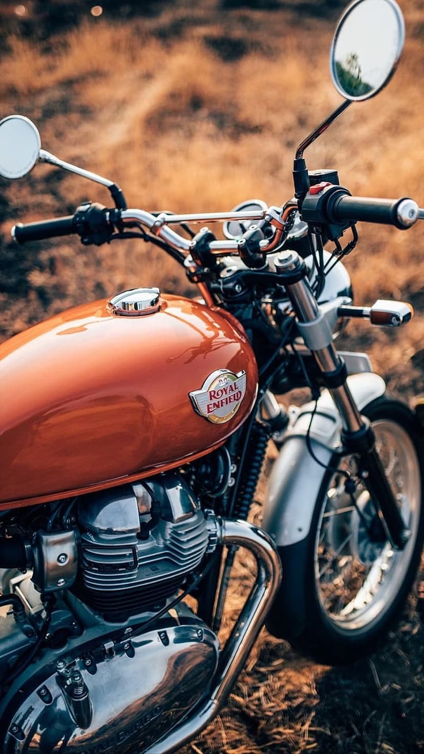 Royal Enfield motorcycle featuring an orange fuel tank and chrome engine parts in a blurred autumnal landscape.