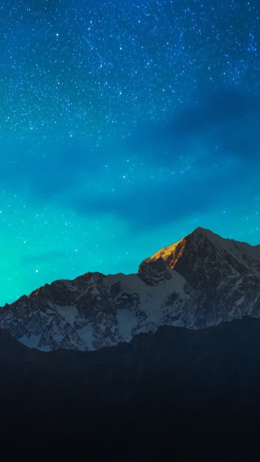 Snow-capped mountain peak glowing under a vibrant blue starry night sky with deep shadows in the foreground.