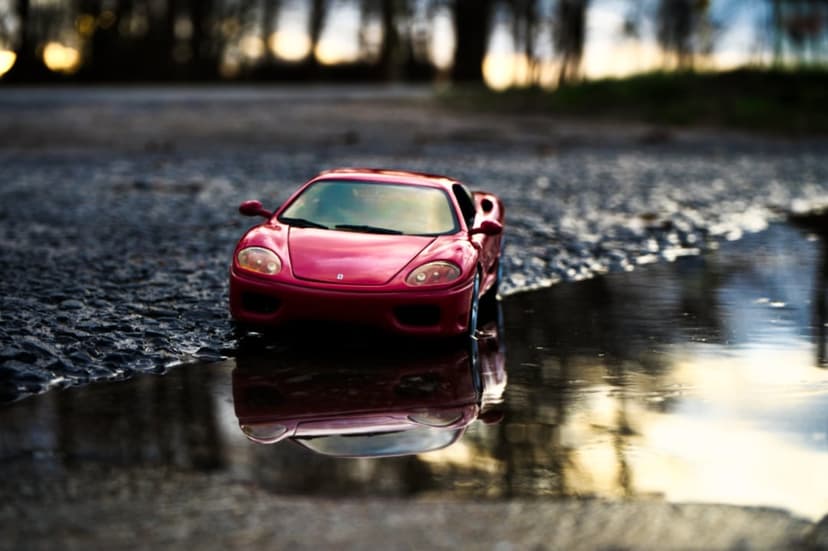 Red miniature sports car reflecting in a shallow water puddle during twilight with a blurry forest background.