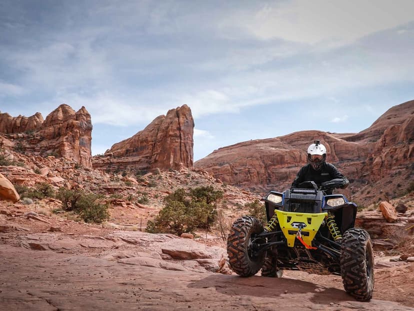 Rider on a blue Polaris ATV climbing a rocky desert slope with giant sandstone formations under a hazy sky.