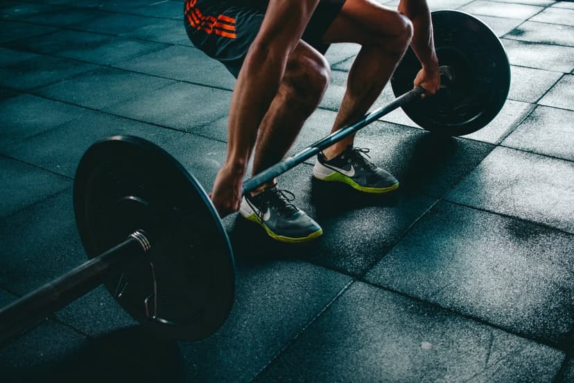 Man's hands and legs positioned at a loaded barbell on a dark rubber gym floor during a strength training session.