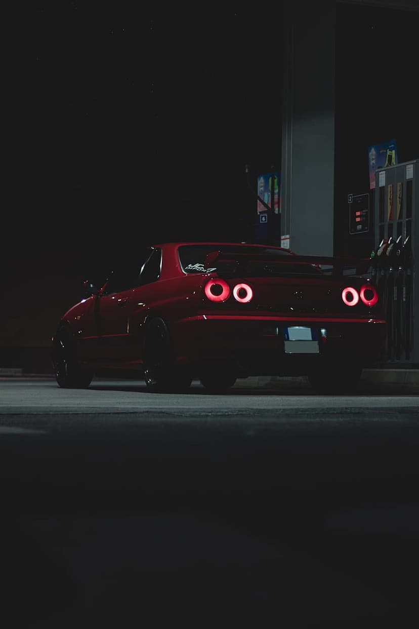 Scarlet supercar with glowing taillights parked at a dark gas station under bright canopy lights at night.