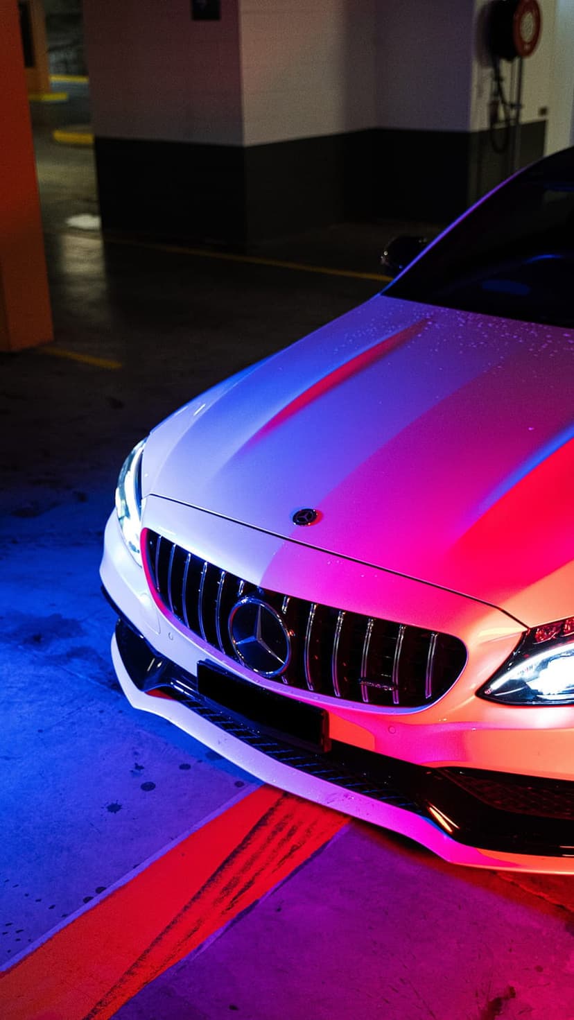 White Mercedes-AMG luxury car with a chrome grille reflects pink and blue neon lights in a dim parking garage.