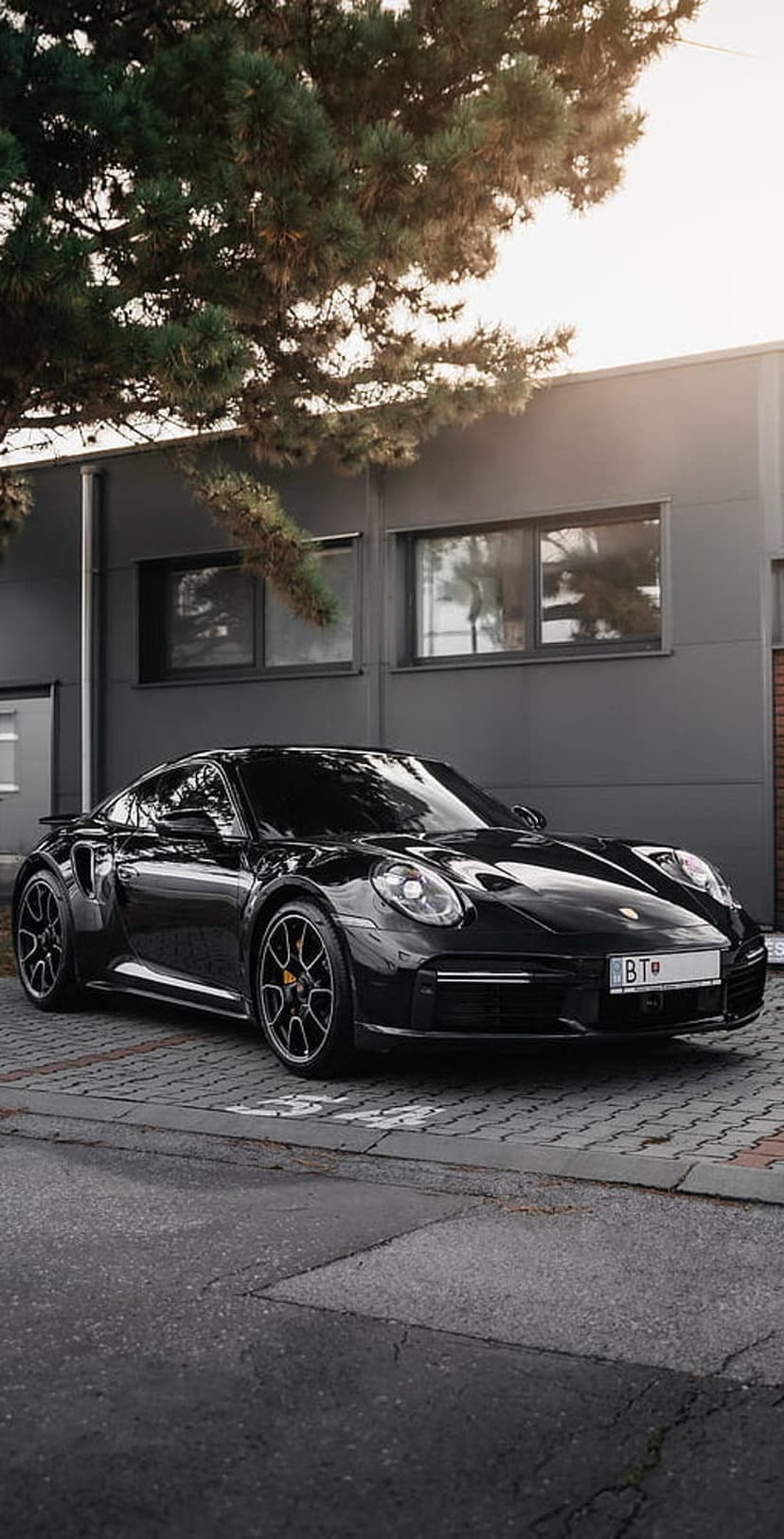 Black Porsche 911 Turbo S parked on an asphalt lot with dappled sunlight and a modern building in the background.