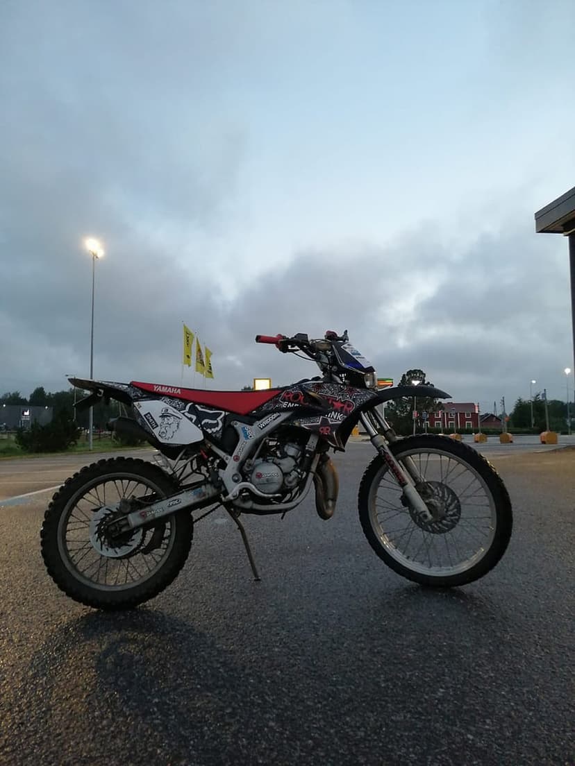 Yamaha dirt bike with a red seat and custom graphics parked on asphalt under a cloudy, overcast sky at dusk.